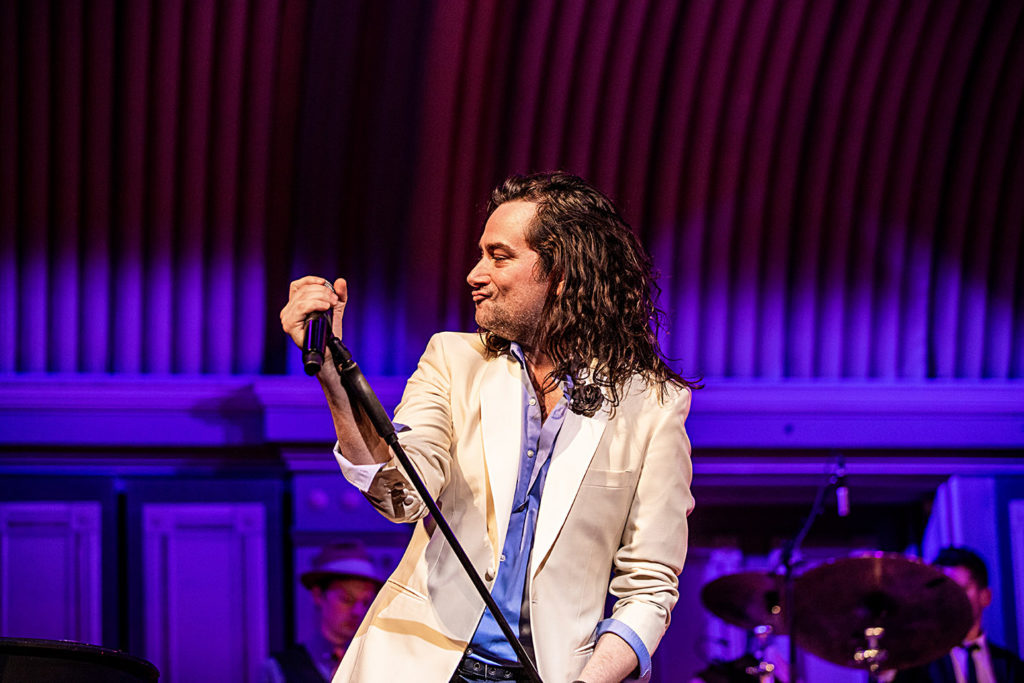 Former American Idol contestant Constantine Maroulis plays the audience with his signature pout. He is holding a microphone. 
